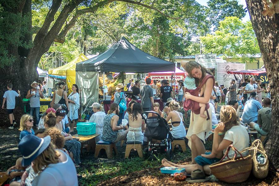 byron-bay-events Crowds enjoying a relaxed outdoor market under shady trees, with food stalls, families, and picnic seating capturing the laid-back atmosphere of byron bay events.